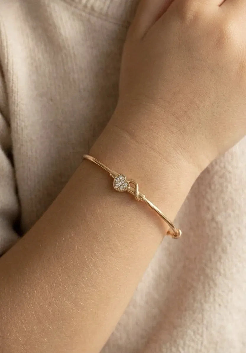 Gold bracelet with a heart-shaped clasp on a wrist against a neutral background