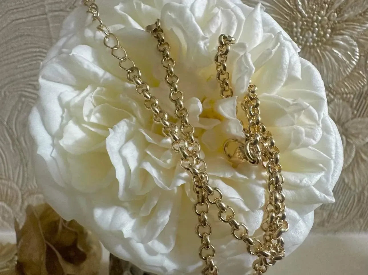 Gold chain necklace draped over a white flower with a textured beige background