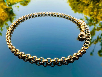 Gold chain bracelet on a reflective surface with blurred greenery in the background