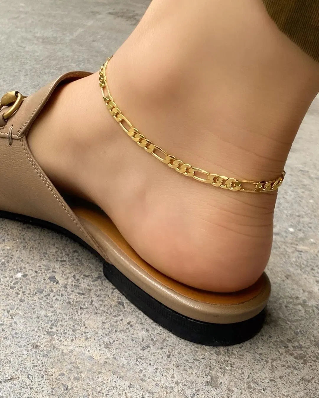 Beige sandal worn on a foot with a gold chain bracelet on a concrete floor.