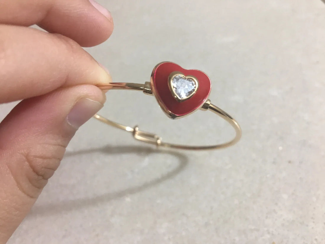 Red heart-shaped bracelet with a gold band held by a hand on a light background