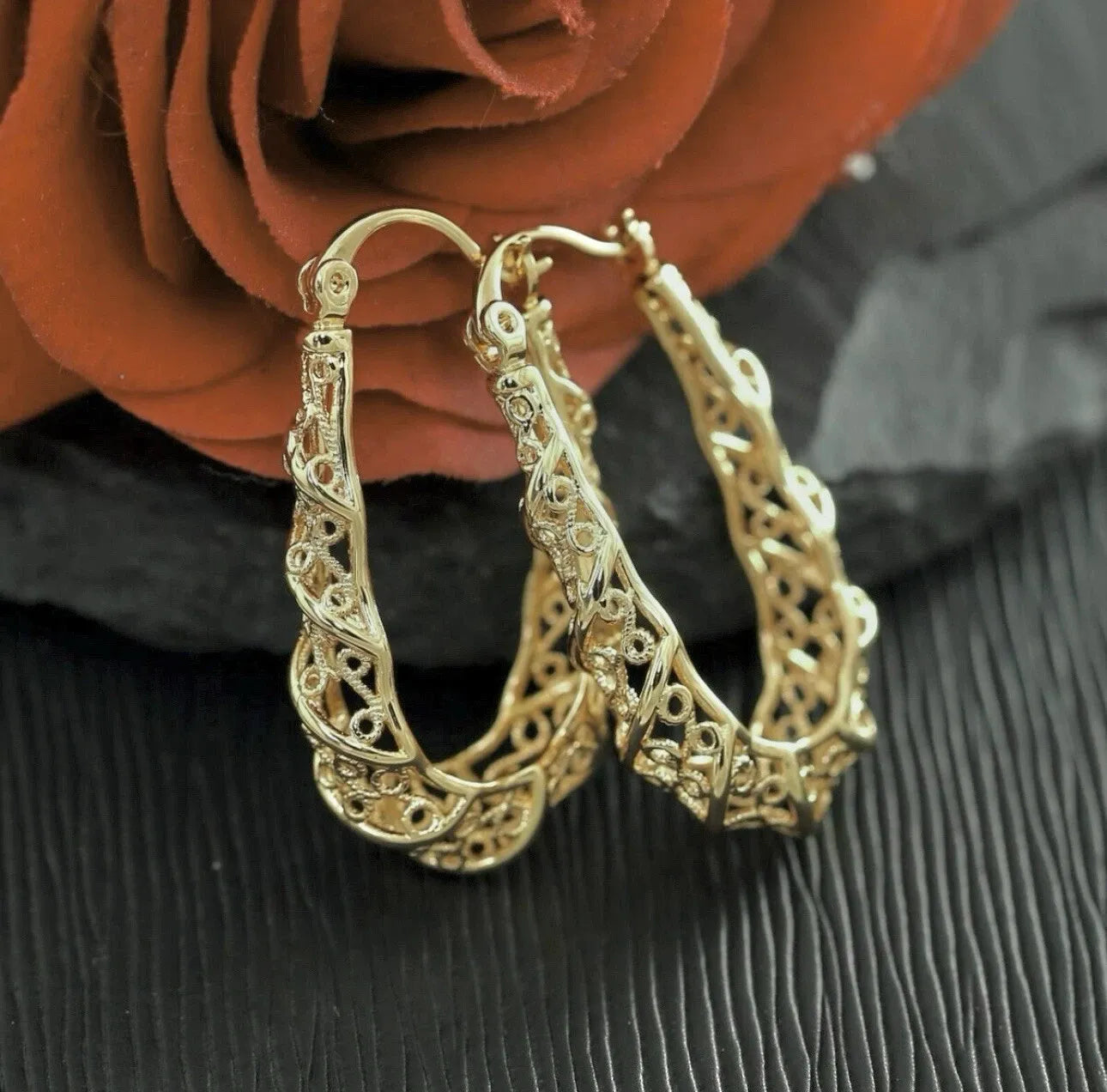 Gold hoop earrings with intricate design on a dark surface with an orange rose in the background