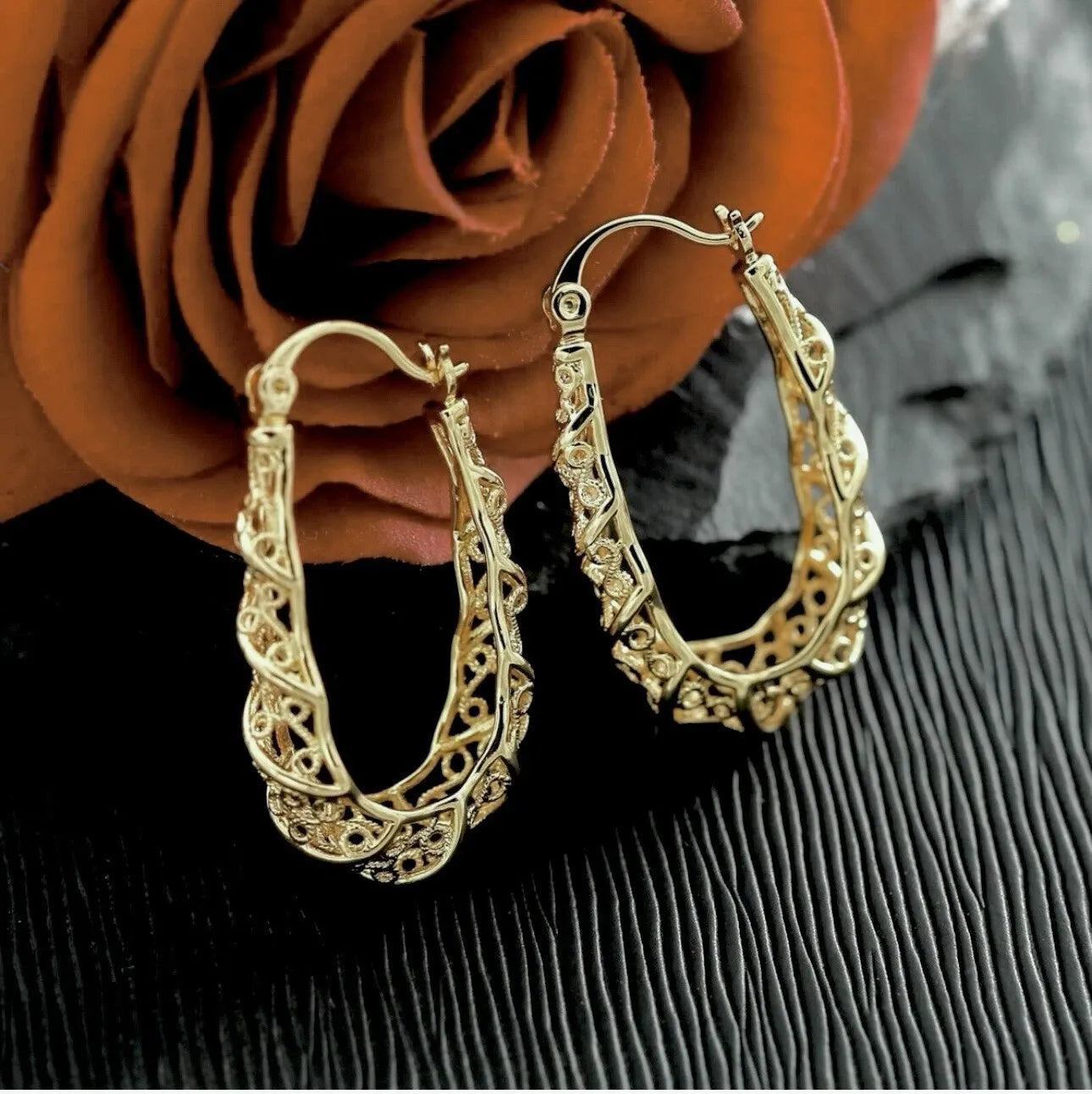 Gold hoop earrings with intricate designs on a textured surface with a rose in the background