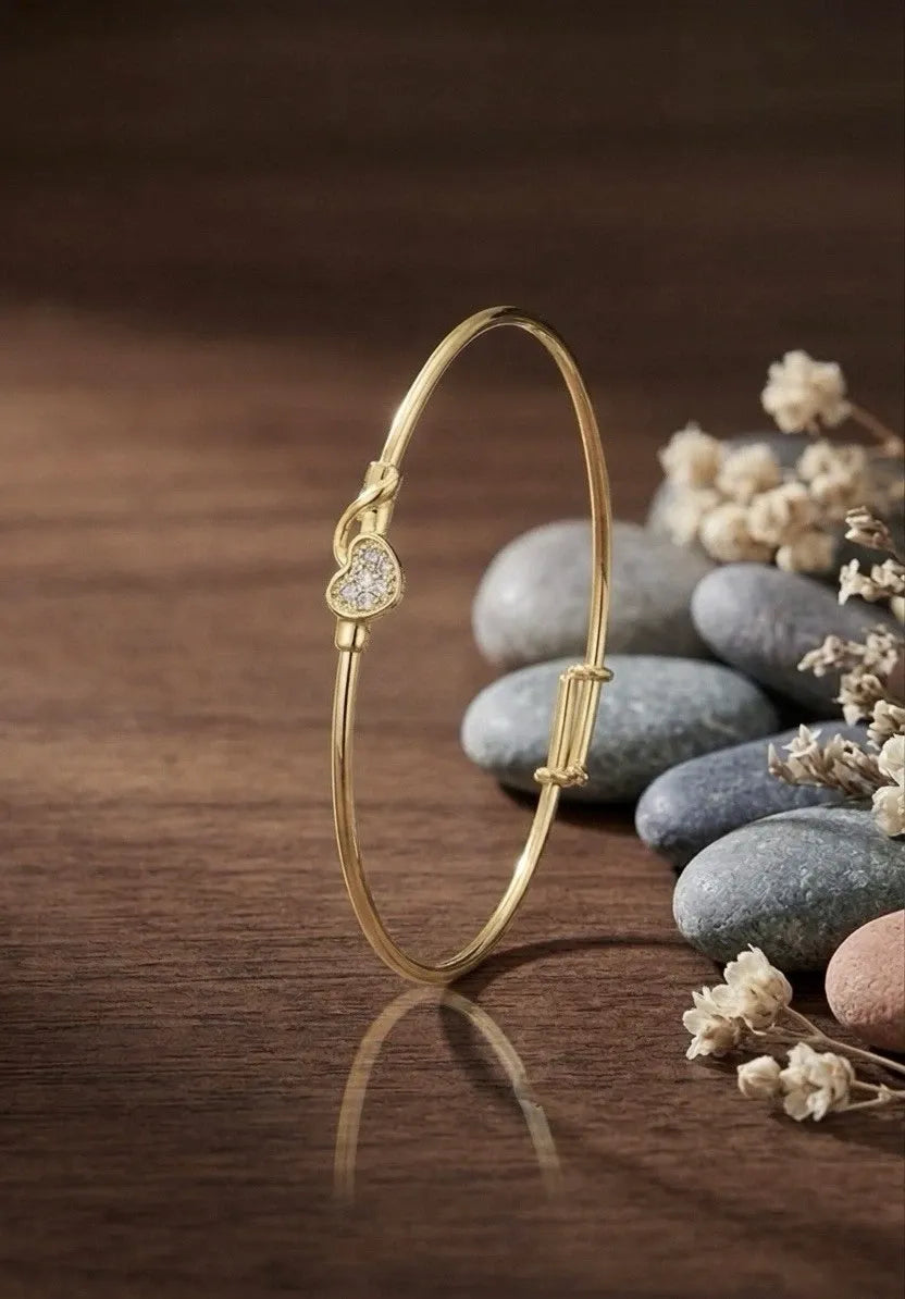 Gold bracelet with a heart-shaped diamond on a wooden surface with stones and flowers.