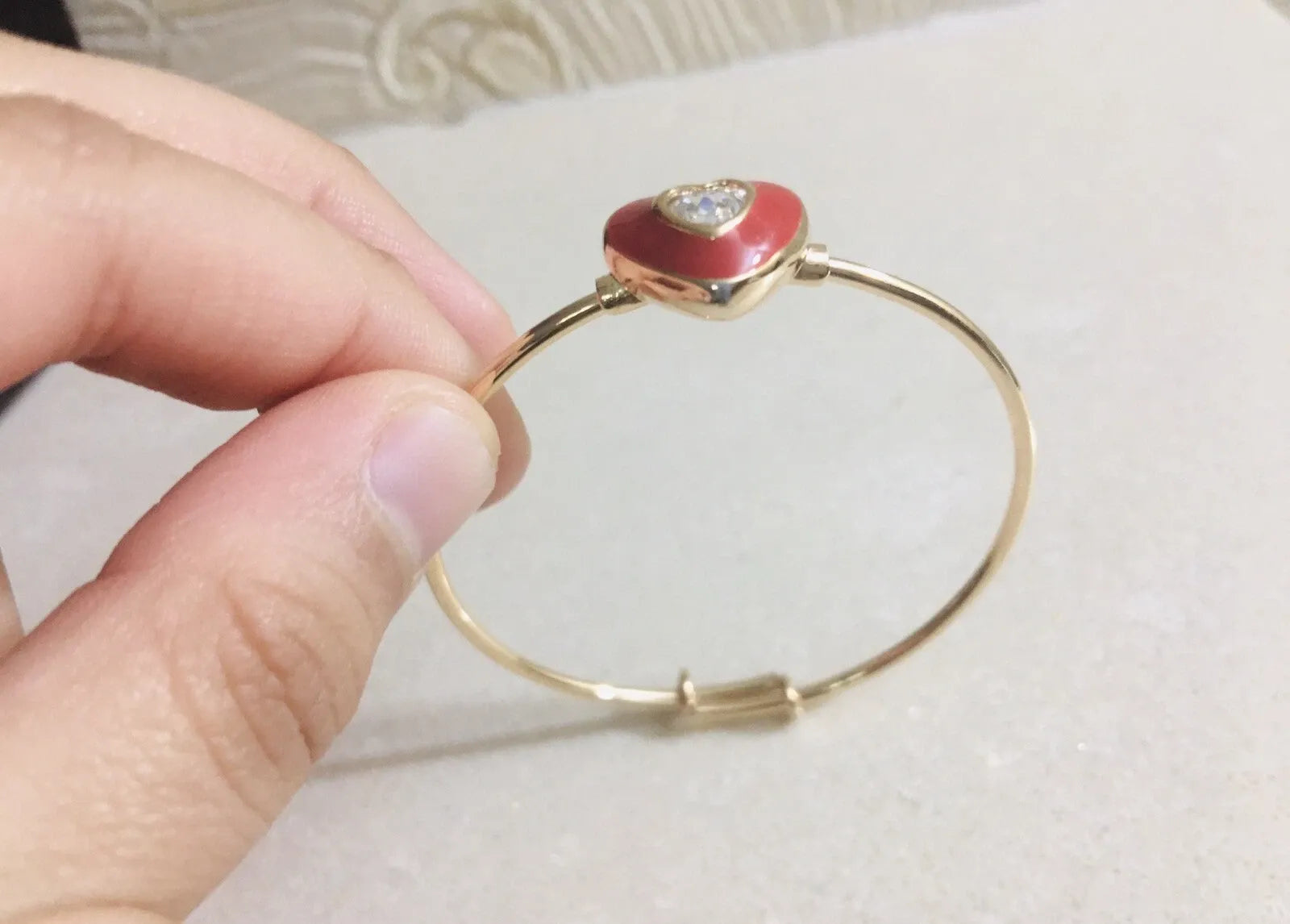 Gold bracelet with a red heart-shaped charm held by a hand on a light background