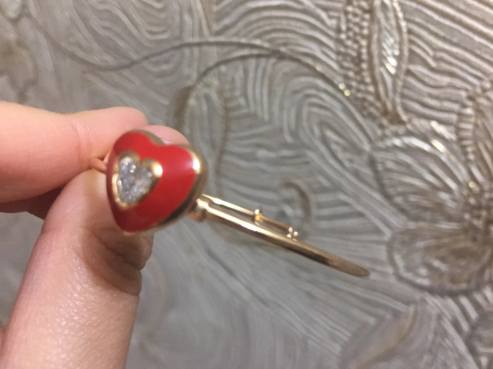Heart-shaped brooch with red enamel and gold detailing held by a hand against a textured background.