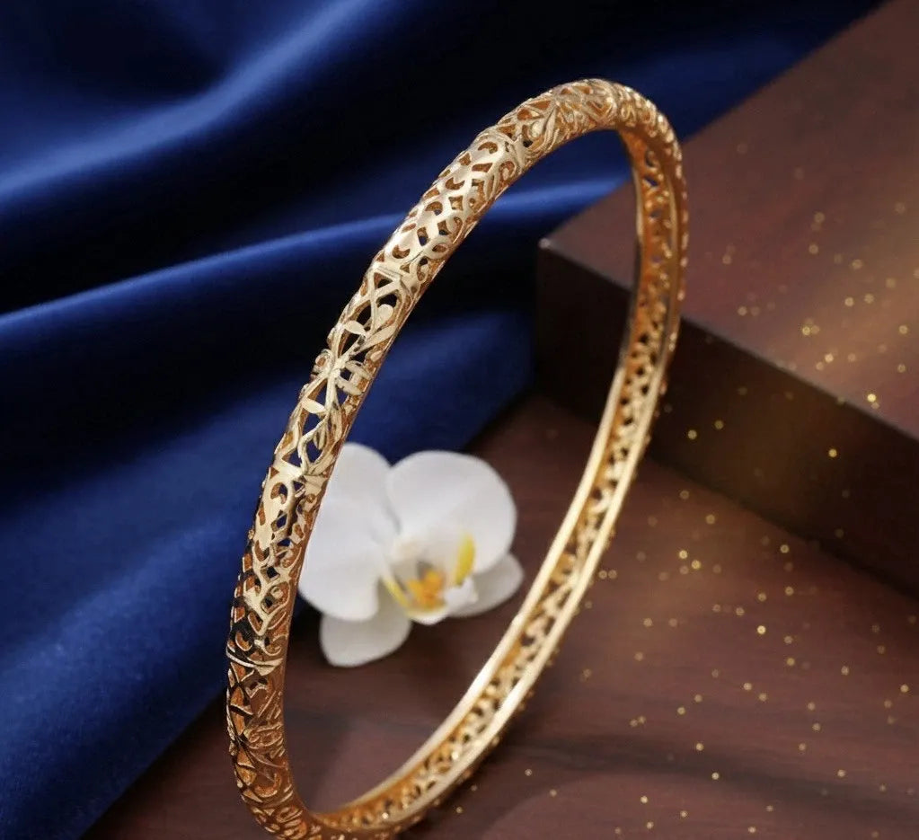 Gold bracelet with intricate design on a dark surface with a white flower.