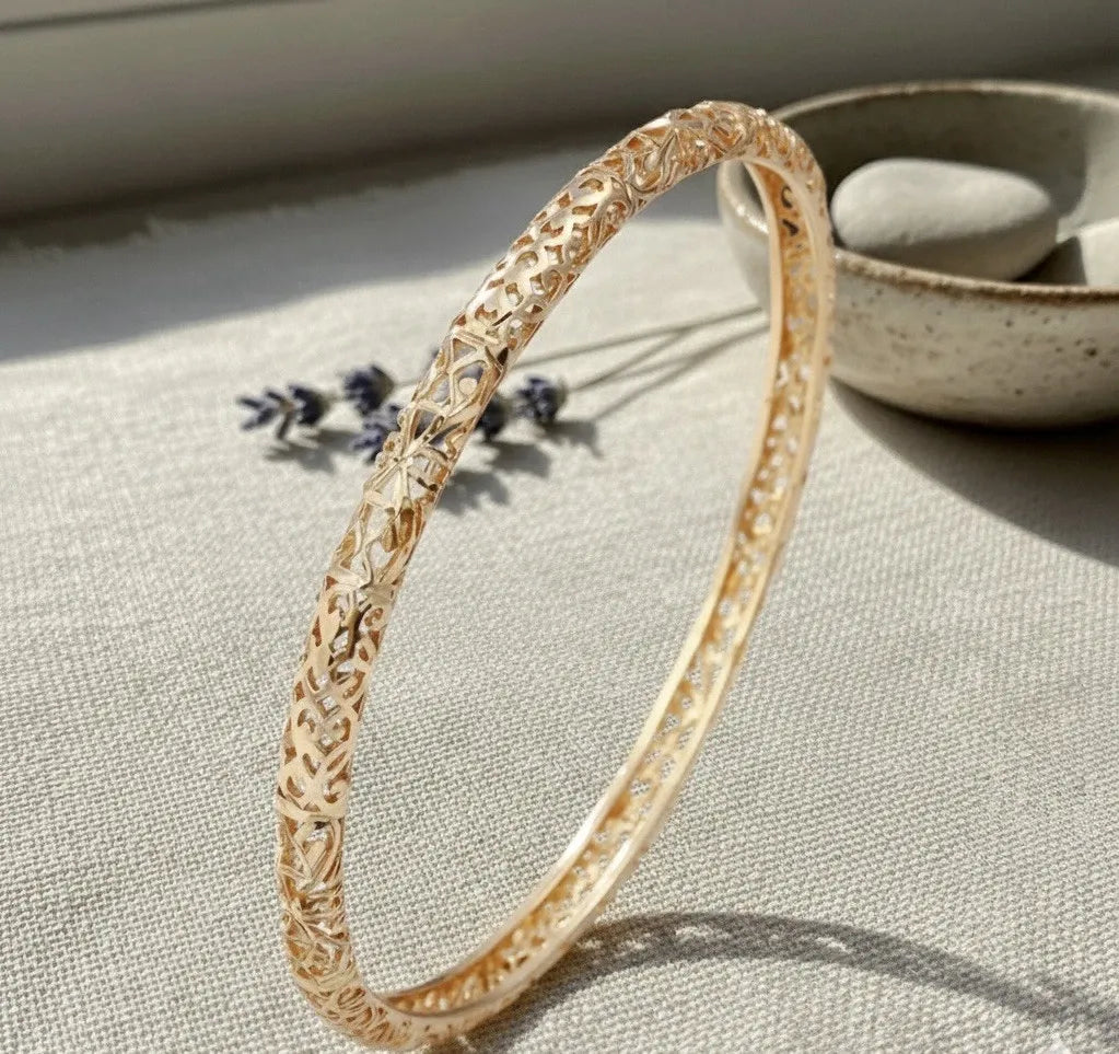 Gold bracelet with intricate design on a textured surface with a bowl and lavender flowers in the background